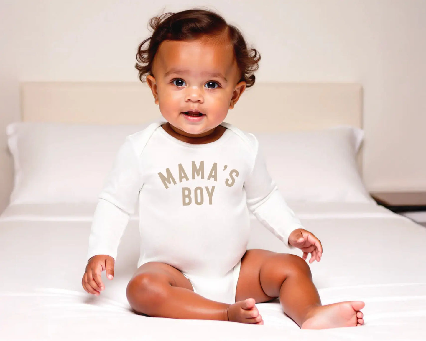 Baby wearing a 'Mama's Boy' onesie sitting on a bed.