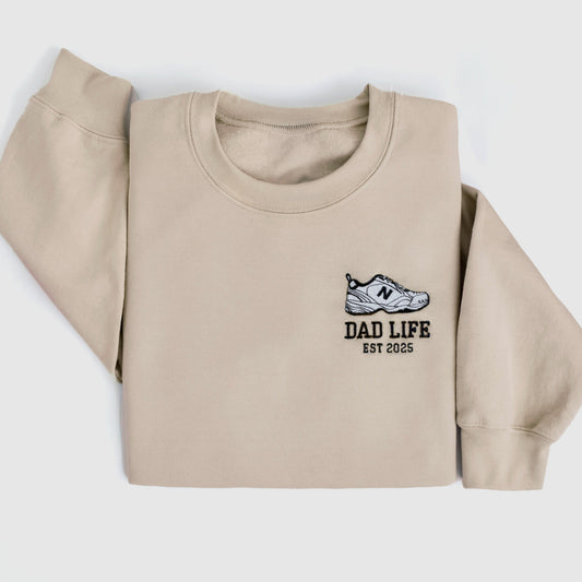 Beige dad life sweatshirt embroidered with new balance dad sneaker and est year. Best gift for new dads close up folded