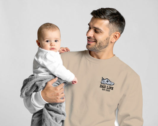 Man holding a baby wearing a beige sweatshirt with 'Dad Life' text on a plain background