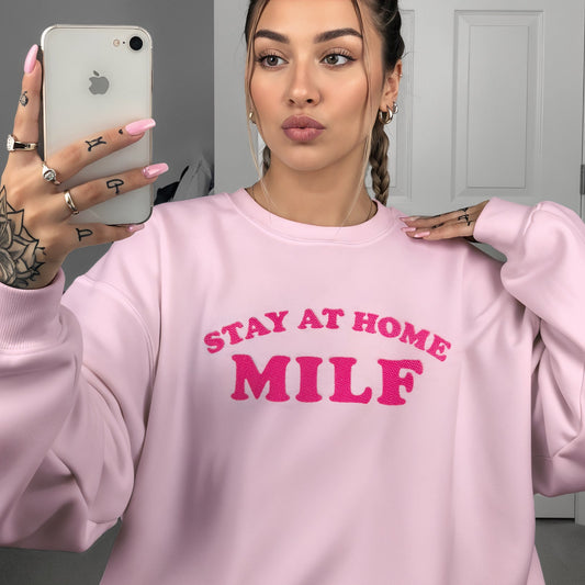 Person wearing a pink sweatshirt with 'STAY AT HOME MILF' text, holding a phone.