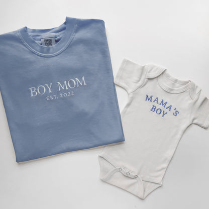 Folded blue shirt with 'BOY MOM EST. 2022' text and a gray baby onesie with 'MAMA'S BOY' text on a light gray background.