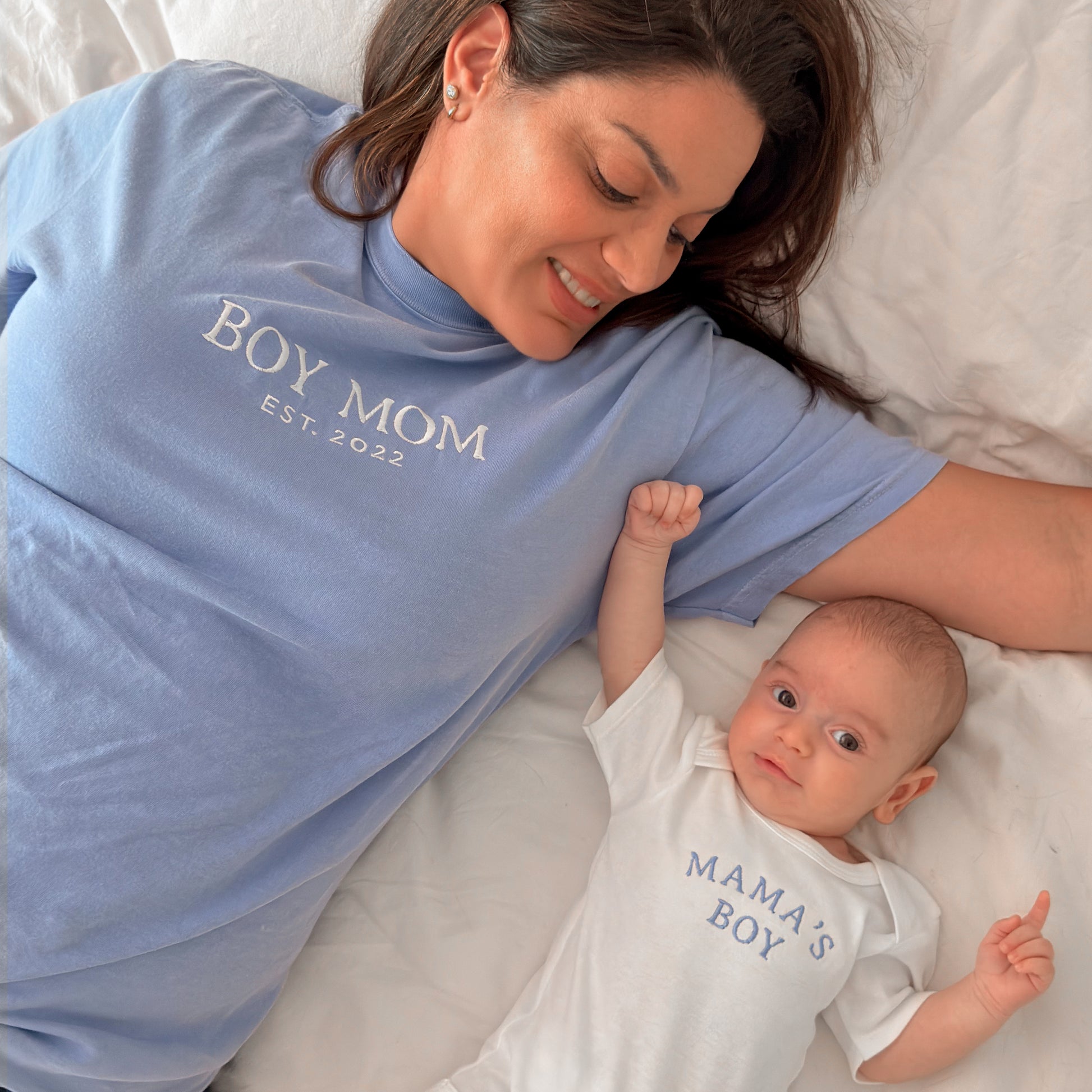 Woman wearing a 'BOY MOM EST. 2022' shirt lying next to a baby wearing a 'MAMA'S BOY' onesie.