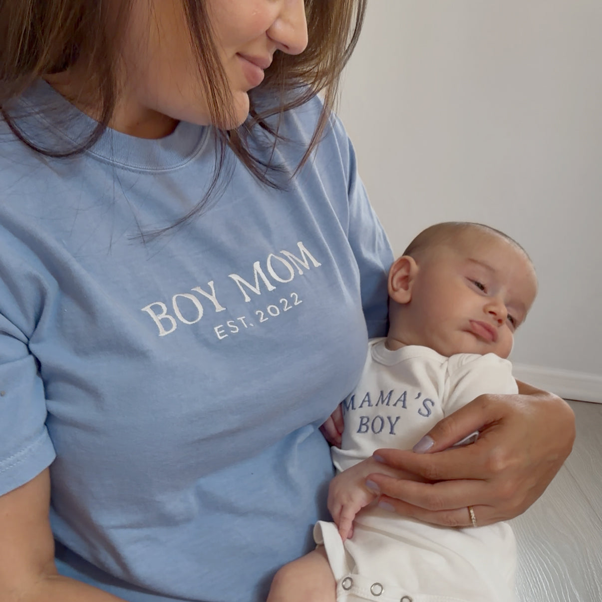 Woman wearing a blue 'BOY MOM EST. 2022' shirt holding a baby in a 'MAMA'S BOY' outfit.
