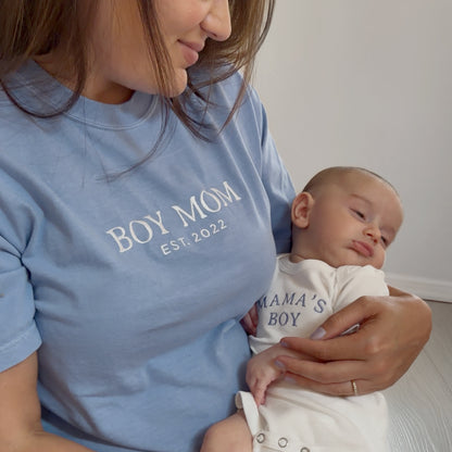 Woman wearing a blue 'BOY MOM EST. 2022' shirt holding a baby in a 'MAMA'S BOY' outfit.