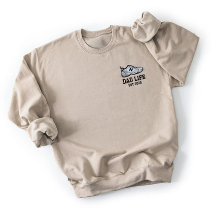 Beige dad life sweatshirt embroidered with new balance dad sneaker and est year. Best gift for new dads flatlay
