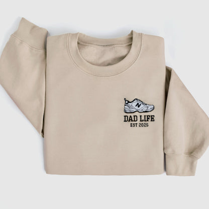 Beige dad life sweatshirt embroidered with new balance dad sneaker and est year. Best gift for new dads close up folded