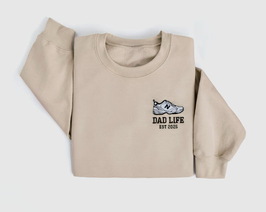 Beige dad life sweatshirt embroidered with new balance dad sneaker and est year. Best gift for new dads close up folded