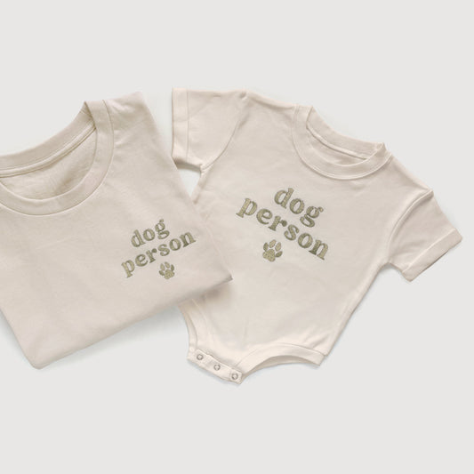 Folded t-shirt and baby onesie with 'dog person' text on a light gray background