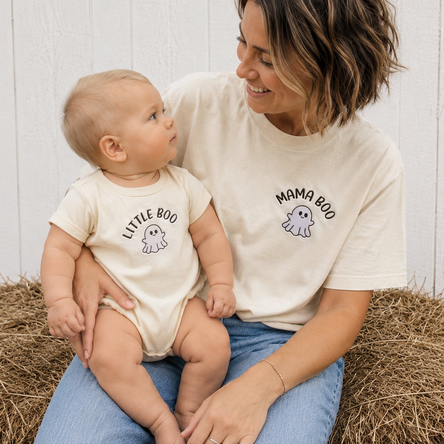 A woman and a baby sit together on a hay bale, wearing matching cream-colored t-shirts. The woman's shirt is labeled 'MAMA BOO' with a ghost graphic, and the baby's shirt is labeled 'LITTLE BOO' with a small ghost graphic.