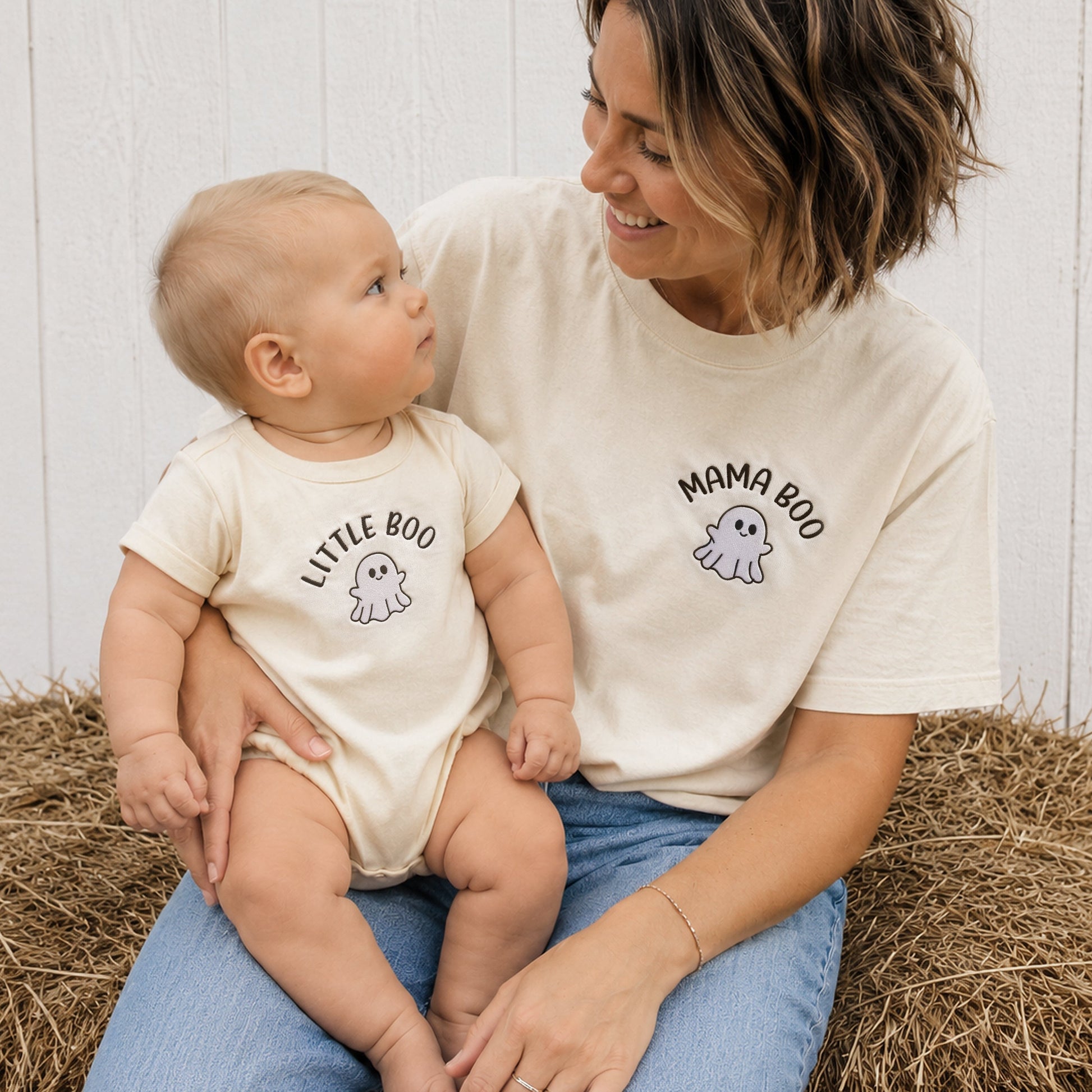 A woman and a baby sit together on a hay bale, wearing matching cream-colored t-shirts. The woman's shirt is labeled 'MAMA BOO' with a ghost graphic, and the baby's shirt is labeled 'LITTLE BOO' with a small ghost graphic.