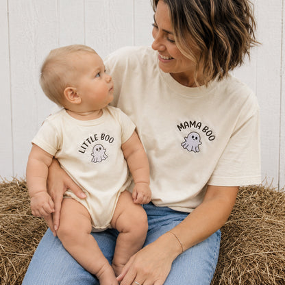A woman and a baby sit together on a hay bale, wearing matching cream-colored t-shirts. The woman's shirt is labeled 'MAMA BOO' with a ghost graphic, and the baby's shirt is labeled 'LITTLE BOO' with a small ghost graphic.