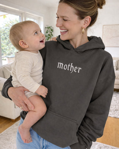 Woman wearing a hoodie with 'mother' printed on it, holding a baby indoors.