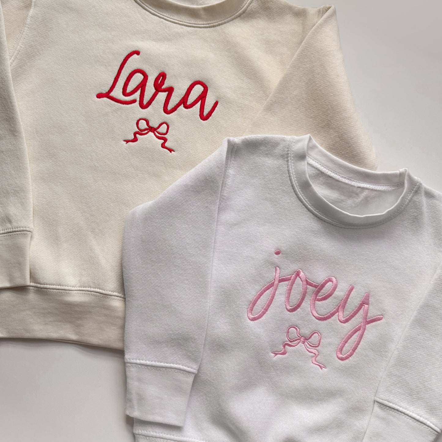 Two sweatshirts, one beige with red text and one white with pink text, on a light gray background.