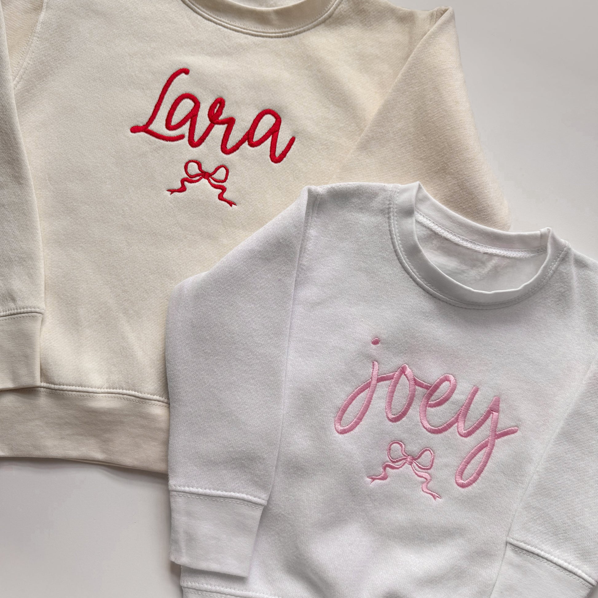 Two sweatshirts, one beige with red text and one white with pink text, on a light gray background.