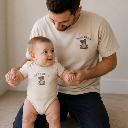 Man wearing a 'Papa Bear' t-shirt and holding hands with a baby wearing a 'Baby Bear' onesie on a carpeted floor.