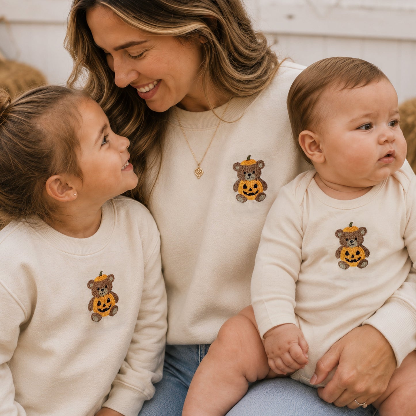 A woman and two children wearing matching cream sweatshirts and bodysuits, each with a small embroidered teddy bear wearing a pumpkin hat. The scene is styled in a warm, autumnal home setting.