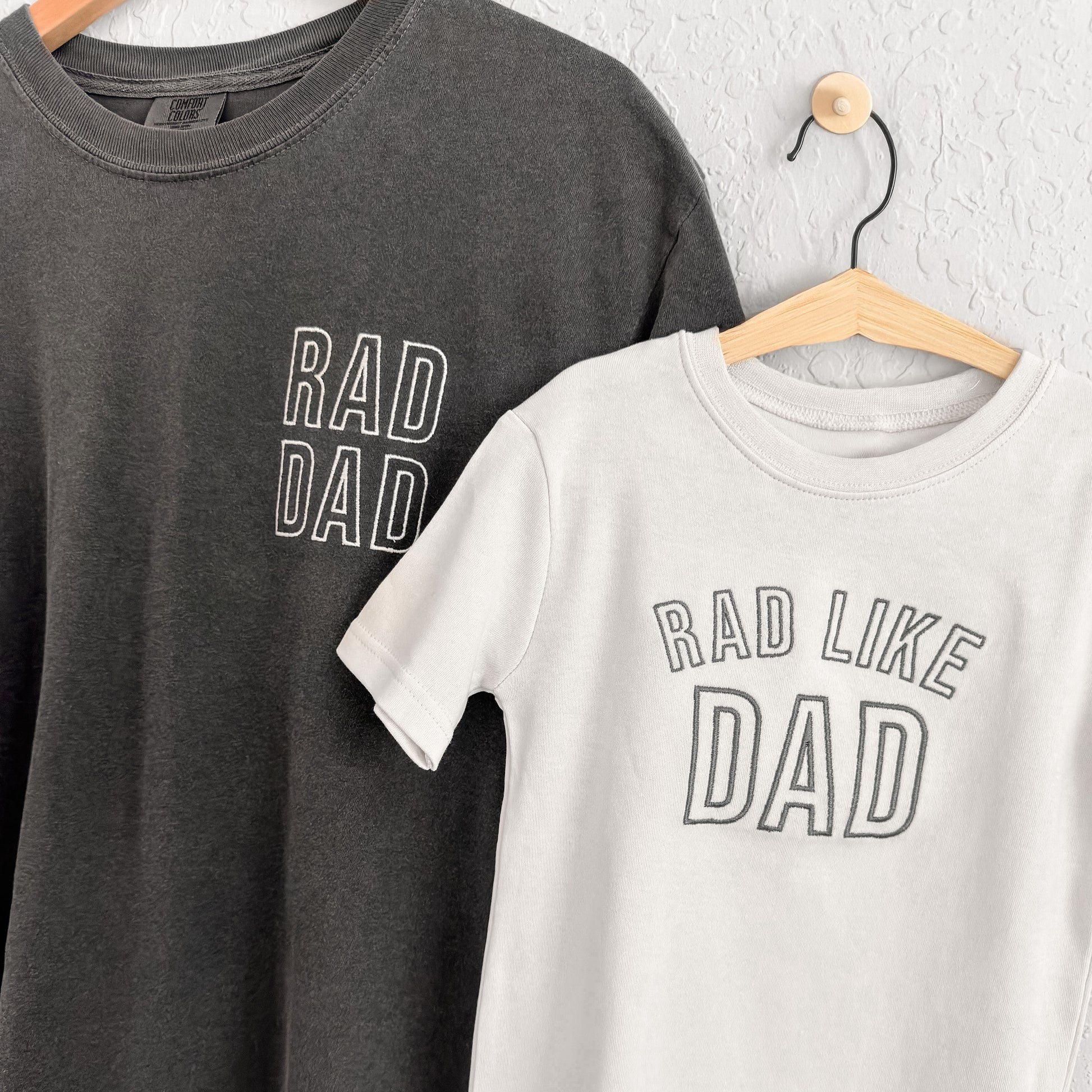 Two t-shirts, one black and one white, with 'RAD DAD' and 'RAD LIKE DAD' text on hangers against a white wall.