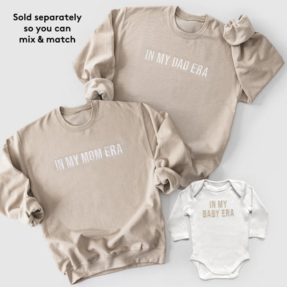 Three beige sweatshirts with text 'IN MY DAD ERA', 'IN MY MOM ERA', and a white baby onesie with 'IN MY BABY ERA' on a light gray background.
